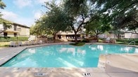 This inviting outdoor amenity space is designed for relaxation and recreation, centered around a generously sized swimming pool with clear, blue water. The pool area is thoughtfully landscaped with mature oak trees that provide natural shade across the concrete surround, creating a comfortable outdoor environment throughout the day. Adjacent to the main pool is a separate hot tub with a decorative stone surround and cascading water feature, adding a resort-like ambiance to the setting.

The patio area includes multiple round tables paired with chairs, each shaded by large green market umbrellas, offering ideal spots for outdoor dining or socializing. A row of lounge chairs is arranged along the exterior wall of the building, perfect for sunbathing or relaxing poolside. An outdoor grill is also available, making the space well-equipped for outdoor gatherings. Wrought iron fencing encloses the pool area, providing a clear boundary while maintaining open sightlines to the surrounding landscaped grounds.

Directly accessible from the pool deck is an interior workspace visible through large floor-to-ceiling windows and sliding glass doors. This bright, modern room features hardwood-style flooring, a conference-style table, ergonomic seating, and recessed lighting, making it a functional complement to the outdoor amenities.