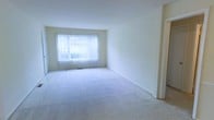 This single-floor space features a spacious, open living area finished with wall-to-wall carpeting in a light neutral tone, contributing to a clean and understated aesthetic throughout. The room is characterized by smooth white walls and a simple, unfussy layout that provides a versatile foundation for a variety of interior styles.

A large window fitted with vertical blinds serves as the focal point of the main living area, allowing generous amounts of natural light to fill the room and creating a bright, airy atmosphere. The window appears to look out toward exterior greenery, offering a pleasant outdoor connection. A floor-level heating vent is positioned beneath the window, indicating a functional climate system integrated into the space.

The overall design is minimal and neutral, with white ceilings that complement the light walls and enhance the sense of openness. A doorway is visible along one wall, suggesting a straightforward and accessible floor plan. The absence of clutter and the abundance of natural light make this space feel welcoming and move-in ready, well-suited for those seeking a clean, adaptable living environment.