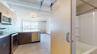 This single-floor unit showcases an industrial-modern aesthetic defined by exposed concrete beam ceilings and polished concrete floors throughout the main living areas. The open-concept kitchen serves as a focal point, featuring a two-toned cabinet design with dark lower cabinetry and light birch upper cabinets, a white countertop peninsula ideal for dining or prep work, and a full suite of stainless steel appliances including a refrigerator, range, microwave, and dishwasher. A sliding barn-style door adds a distinctive architectural touch near the kitchen entry.

The bedroom areas feature patterned carpet flooring, built-in closet storage with birch panel doors, and windows that draw in natural light while offering exterior views. Exposed conduit and structural beams lend an authentic loft character to these rooms.

The bathroom is cleanly designed with polished concrete flooring, a tub-shower combination, dark wood vanity cabinetry, and chrome fixtures and grab bars. Throughout the unit, the interplay of raw industrial elements—exposed ceilings, visible piping—and modern finishes creates a cohesive, contemporary living environment with a distinctive urban character.