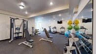 This fitness center offers a generously sized, single-level workout space designed to accommodate a full range of exercise activities. The layout is open and functional, featuring durable rubber flooring throughout that provides both safety and comfort during workouts. Recessed lighting and ceiling fans ensure the space remains well-lit and well-ventilated, while a wall-mounted flat-screen TV adds entertainment during cardio sessions.

The cardio zone is equipped with multiple treadmills, an elliptical trainer, a stationary bike, and a rowing machine, providing ample options for aerobic exercise. The strength training area includes a multi-function cable machine, adjustable weight benches, a comprehensive dumbbell rack, and a selection of medicine balls and stability balls neatly organized on a dedicated rack.

Mirrored wall panels enhance the sense of space and allow users to monitor their form during workouts. A split-unit air conditioning system maintains a comfortable temperature year-round. Natural light filters in through large windows and a set of French doors, which open to an outdoor area, lending the space a bright, airy ambiance. The neutral, light-toned walls and clean design create a focused and motivating environment suited for a variety of fitness routines.