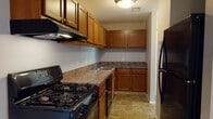 The Spruce is a thoughtfully laid-out 2-bedroom, 1-bathroom single-floor residence designed to offer comfortable, functional living. The kitchen is a standout feature, outfitted with warm honey-toned cabinetry providing generous upper and lower storage, stone-patterned laminate countertops, and a full suite of black appliances including a gas range, dishwasher, and refrigerator. Stone-look tile flooring completes the kitchen's practical and cohesive aesthetic. The open living and dining area features plush carpeting and a built-in shelving unit, offering both comfort and convenient storage. A pendant light fixture adds a subtle design touch, while a sliding glass door leads to views of a grassy outdoor area, bringing in natural light. Both bedrooms are carpeted and feature large windows that frame outdoor green space views, creating a bright and airy atmosphere. The full bathroom includes a tiled tub-shower combination, a wood-toned vanity with stone-pattern countertop, and matching tile flooring consistent with the kitchen. Throughout the residence, neutral wall tones and consistent finishes create a clean, cohesive environment well-suited for everyday living.