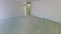 This single-floor space presents a straightforward, functional layout centered around an open living area. The room is finished with wall-to-wall carpeting in a neutral, light gray-green tone that extends throughout, providing a soft and cohesive foundation underfoot. The walls are painted in a clean, off-white hue, contributing to a bright and understated aesthetic.

A floor-level air vent is visible along the base of one wall, and a baseboard heating unit runs along the far wall, indicating a practical heating setup suited for year-round comfort. A structural column or partial wall protrudes slightly into the space, adding subtle architectural definition to the room's otherwise clean lines.

An interior doorway leads into an adjoining area, which appears to be a secondary room or hallway illuminated by overhead lighting, suggesting additional functional space beyond the main living area. The overall ambiance is clean, minimal, and move-in ready, with neutral finishes that offer flexibility for a variety of furnishing styles. The space conveys a practical design ethos, prioritizing utility and ease of use throughout its layout.