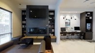The Cyber Lounge is a single-level space that seamlessly blends comfort and functionality through a contemporary design aesthetic. The focal point of the lounge is a bold, dark-tiled fireplace wall, which extends floor to ceiling and is flanked by built-in bookshelves filled with books and decorative accents. A large flat-screen television is mounted prominently above the fireplace, creating an inviting entertainment hub. In front of the fireplace, low-profile bench seating with warm-toned cushions provides a relaxed gathering spot, complemented by a dark coffee table and leather seating arranged on a patterned area rug. The light hardwood flooring contrasts elegantly with the dark feature wall, adding warmth and balance to the space. Adjacent to the lounge area, a dedicated workstation zone offers desktop computers, a printer, and task lighting mounted on adjustable wall-mounted arms, making it well-equipped for productivity needs. Recessed ceiling lighting throughout ensures the space remains bright and welcoming. The overall ambiance is modern and polished, combining a cozy lounge atmosphere with practical business amenities.