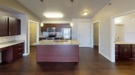 This single-floor residence is defined by its striking vaulted ceilings, which lend an expansive, airy quality to the open-concept living and kitchen area. Rich, dark hardwood flooring runs throughout the main living spaces, complementing the warm neutral tones of the walls and creating a cohesive, polished aesthetic.

The kitchen is a standout feature, equipped with a large center island topped with granite countertops and an undermount sink. Dark espresso cabinetry provides ample storage, while a full suite of stainless steel appliances—including a refrigerator, range, and microwave—adds a modern, functional touch. Pendant lighting above the island enhances the kitchen's inviting atmosphere. An adjacent walk-in pantry offers generous storage space.

The open living area flows seamlessly from the kitchen, benefiting from the soaring vaulted ceiling and access to an entry door with a sidelight window. The bathroom features granite countertops, a dark wood vanity with brushed nickel hardware, and a large mirror with contemporary lighting above.

Bedrooms are carpeted for comfort, with windows that allow natural light to fill the rooms. A dedicated laundry room houses a full-sized washer and dryer with overhead shelving for added convenience, completing this well-appointed, single-level home.