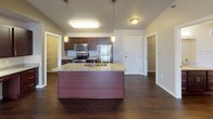 This single-floor residence is defined by its striking vaulted ceilings, which lend an expansive, airy quality to the open-concept living and kitchen area. Rich, dark hardwood flooring runs throughout the main living spaces, complementing the warm neutral tones of the walls and creating a cohesive, polished aesthetic.

The kitchen is a standout feature, equipped with a large center island topped with granite countertops and an undermount sink. Dark espresso cabinetry provides ample storage, while a full suite of stainless steel appliances—including a refrigerator, range, and microwave—adds a modern, functional touch. Pendant lighting above the island enhances the kitchen's inviting atmosphere. An adjacent walk-in pantry offers generous storage space.

The open living area flows seamlessly from the kitchen, benefiting from the soaring vaulted ceiling and access to an entry door with a sidelight window. The bathroom features granite countertops, a dark wood vanity with brushed nickel hardware, and a large mirror with contemporary lighting above.

Bedrooms are carpeted for comfort, with windows that allow natural light to fill the rooms. A dedicated laundry room houses a full-sized washer and dryer with overhead shelving for added convenience, completing this well-appointed, single-level home.