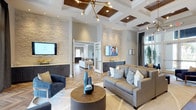 This beautifully designed single-floor space exudes a refined, contemporary aesthetic that is both welcoming and visually impressive. The open layout is anchored by striking coffered ceilings with warm metallic inlays, complemented by a statement chandelier with brass and gold accents that adds a touch of glamour to the interior. Underfoot, herringbone-patterned hardwood flooring runs throughout, lending a sense of timeless elegance and cohesion to the space.

Natural stacked stone accent walls provide rich textural contrast against the soft, neutral color palette of grays and whites, creating a sophisticated backdrop for the carefully curated furnishings. The main seating area features plush gray sofas and deep navy accent chairs arranged around a circular wood coffee table, offering a comfortable and stylish gathering space. Multiple flat-screen displays are thoughtfully integrated into the design, enhancing the functionality of the room.

Dark wood cabinetry lines portions of the walls, providing ample storage while contributing to the layered, upscale ambiance. Large windows dressed with floor-length drapes invite natural light into the space, while glimpses of lush greenery add a refreshing organic element. A connected corridor leads to additional areas, suggesting a spacious and well-appointed floor plan throughout.