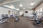 Located at 1401 College Ave in Manhattan, Kansas, this well-equipped fitness center offers a comprehensive workout environment spread across a single floor. The space is divided into distinct functional zones, each designed to support a variety of exercise routines. The main cardio area features multiple treadmills, an elliptical trainer, a stationary air bike, and a rowing machine, all arranged along a row of large windows that overlook an outdoor pool area, filling the room with natural light. A dedicated strength training section houses a cable cross machine, an adjustable weight bench, and a full rack of dumbbells in varying weights, complemented by floor-to-ceiling mirrors that enhance the sense of space and allow for proper form monitoring. An additional area provides stability balls, resistance bands, and exercise mats for flexibility and functional fitness work. The flooring throughout is durable rubber, well-suited for high-traffic athletic use. The interior is finished in neutral gray tones with bright overhead fluorescent lighting ensuring excellent visibility at all hours. A water cooler and a wall-mounted flat-screen television are also present, adding convenience and comfort. A secondary entry with glass-panel doors connects to an adjacent facility, further enhancing accessibility within the broader complex.
