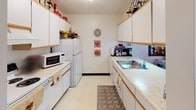 This single-floor residence offers a thoughtfully organized layout with multiple rooms connected by carpeted hallways and consistent neutral finishes throughout. The living room is bright and inviting, featuring large sliding glass doors with vertical blinds that allow ample natural light to fill the space, complemented by textured ceilings and baseboard heating. The kitchen is a galley-style design equipped with white cabinetry, light countertops, a full suite of appliances including a refrigerator, stove, and microwave, along with a double-basin sink. Adjacent to the kitchen, the dining area accommodates a full dining table and chairs beneath a ceiling fan, with a decorative mirror adding depth to the room. The primary bedroom is generously sized, featuring a ceiling fan, a classic four-poster bed frame, plush carpet, and a large dresser with mirror. A second bedroom offers closet storage, a window with sheer curtains providing soft natural light, and comfortable proportions suitable for various uses. A third, cozier bedroom includes its own closet and direct hallway access. The bathroom features a wide vanity with a light countertop, an illuminated mirror, and bright overhead lighting. Throughout the residence, warm wood-toned trim and soft carpet flooring create a cohesive and comfortable ambiance.