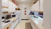 This single-floor residence offers a thoughtfully organized layout with multiple rooms connected by carpeted hallways and consistent neutral finishes throughout. The living room is bright and inviting, featuring large sliding glass doors with vertical blinds that allow ample natural light to fill the space, complemented by textured ceilings and baseboard heating. The kitchen is a galley-style design equipped with white cabinetry, light countertops, a full suite of appliances including a refrigerator, stove, and microwave, along with a double-basin sink. Adjacent to the kitchen, the dining area accommodates a full dining table and chairs beneath a ceiling fan, with a decorative mirror adding depth to the room. The primary bedroom is generously sized, featuring a ceiling fan, a classic four-poster bed frame, plush carpet, and a large dresser with mirror. A second bedroom offers closet storage, a window with sheer curtains providing soft natural light, and comfortable proportions suitable for various uses. A third, cozier bedroom includes its own closet and direct hallway access. The bathroom features a wide vanity with a light countertop, an illuminated mirror, and bright overhead lighting. Throughout the residence, warm wood-toned trim and soft carpet flooring create a cohesive and comfortable ambiance.