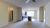 This single-floor residence showcases an open-concept design that connects the living area and kitchen in one cohesive space. The living area is finished with plush carpeting and features a ceiling fan overhead, contributing to a comfortable and functional atmosphere. A smooth transition leads into the kitchen, where the flooring shifts to hard surface material, clearly delineating the two spaces.

The kitchen is well-appointed with dark cabinetry, stainless steel appliances including a refrigerator and oven range, and a central island with a sink and pendant lighting above. Track lighting runs along the kitchen ceiling, providing focused task illumination. A large window above the sink area allows natural light to fill the space, brightening the neutral-toned walls throughout.

A hallway visible to the left leads to additional areas of the unit, suggesting a thoughtful layout with clear separation between living and private spaces. The overall design balances modern finishes with a neutral, move-in-ready palette, making it a versatile and practical space suited to a variety of lifestyles.