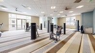 This well-equipped fitness center at 102 N Lemon Ave in Ontario, California, offers a generously proportioned single-floor layout designed to accommodate a comprehensive range of workout activities. The open floor plan is anchored by a distinctive striped carpet in neutral tones of beige, tan, and brown, providing a clean and organized visual flow throughout the space.

The facility is outfitted with an extensive selection of strength training machines, including cable systems, cable crossover units, and a variety of selectorized resistance machines positioned throughout the floor. The cardio section features multiple treadmills, elliptical trainers, and stationary bikes, all conveniently arranged along windowed walls that allow natural light to fill the space and offer views of the surrounding outdoor greenery.

The ceiling features a suspended tile grid with integrated recessed lighting supplemented by multiple ceiling fans, ensuring the space remains well-lit and comfortable during use. A staircase with iron railings and integrated parallel bars—suitable for rehabilitation or mobility exercises—serves as a notable architectural feature within the center.

Accessibility is supported by an elevator on the premises, and the space maintains a clean, professional atmosphere with soft blue accent walls complementing the neutral beige tones. Posted fitness center guidelines reflect a well-managed, community-oriented facility.