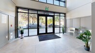 Located at 2578 James St in Abbotsford, BC, this building lobby serves as the primary entrance and common area for residents. The space is defined by its impressive double-height ceiling and a striking floor-to-ceiling arched glass facade at the main entrance, flooding the interior with abundant natural light and offering clear sightlines to the landscaped exterior. Large-format neutral tile flooring extends throughout, lending a clean and polished aesthetic to the entire area.

The entrance vestibule features black-framed glass double doors bearing the building's address and Parkview branding, complemented by decorative potted greenery and a seating area. A dark entry mat anchors the threshold neatly.

Beyond the vestibule, the hallway is lined with panelled grey wainscoting that adds a refined, contemporary character. Secured interior doors with key-fob access panels lead residents further into the building. Safety features including a fire safety plan display, emergency signage, and security cameras are prominently integrated throughout.

A dedicated mailroom alcove houses a full bank of individual stainless steel mailboxes, a tenant notice board, and a parcel drop box, providing convenient mail management for all residents. The overall ambiance is modern, well-lit, and functional — presenting a welcoming and secure point of entry.