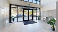 Located at 2578 James St in Abbotsford, BC, this building lobby serves as the primary entrance and common area for residents. The space is defined by its impressive double-height ceiling and a striking floor-to-ceiling arched glass facade at the main entrance, flooding the interior with abundant natural light and offering clear sightlines to the landscaped exterior. Large-format neutral tile flooring extends throughout, lending a clean and polished aesthetic to the entire area.

The entrance vestibule features black-framed glass double doors bearing the building's address and Parkview branding, complemented by decorative potted greenery and a seating area. A dark entry mat anchors the threshold neatly.

Beyond the vestibule, the hallway is lined with panelled grey wainscoting that adds a refined, contemporary character. Secured interior doors with key-fob access panels lead residents further into the building. Safety features including a fire safety plan display, emergency signage, and security cameras are prominently integrated throughout.

A dedicated mailroom alcove houses a full bank of individual stainless steel mailboxes, a tenant notice board, and a parcel drop box, providing convenient mail management for all residents. The overall ambiance is modern, well-lit, and functional — presenting a welcoming and secure point of entry.