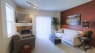 This single-floor living space centers around a well-appointed living room designed for both comfort and style. A tile-surround fireplace serves as a focal point on one wall, topped by a mounted flat-screen television, creating a functional and inviting entertainment area. A striking deep red accent wall adds warmth and visual depth to the room, complemented by a colorful landscape artwork that enhances the overall aesthetic.

The space is finished with plush wall-to-wall carpeting underfoot, contributing to a warm and comfortable atmosphere. A large sliding glass door dressed with vertical blinds allows abundant natural light to fill the room while providing access to the outdoors. Track lighting on the ceiling offers adjustable illumination throughout the space.

Seating is arranged around a sleek glass-top coffee table, with a white tufted sofa and a patterned accent chair providing ample seating options. A tall indoor plant positioned near the sliding door adds a natural, refreshing element. Overall, the space balances practical features with decorative touches to create a welcoming and livable environment.