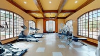 Located in Humble, TX, this fitness center offers a thoughtfully designed exercise space that combines functionality with impressive architectural detail. The room is defined by its striking coffered ceiling with rich dark wood beams, warm golden-toned walls, and a series of large arched windows with wood-framed grid panes that line the perimeter, flooding the space with natural light and offering views of the outdoor surroundings, including a pool area. Ceiling fans integrated into the coffered structure provide additional comfort during workouts. The flooring features a durable speckled tile pattern with lighter inset squares, creating a clean and practical surface suited for high-traffic use. The exercise room is well-stocked with a comprehensive selection of equipment, including multiple treadmills, elliptical trainers, a stationary cycle with an integrated display, and a rowing-style machine. Strength training is supported by a Hoist multi-station cable machine, a weight bench, and a freestanding dumbbell rack with a full range of weights. Stability balls and exercise mats round out the available equipment. Double wood entry doors provide a grand entrance, and informational signage is posted near the entry area. The overall ambiance is bright, open, and inviting, making this fitness center a well-appointed facility for a complete workout experience.