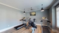 Located in Daphne, Alabama, this single-floor fitness center offers a clean, well-appointed space designed for effective and comfortable workouts. The room is finished with soft gray walls, crisp white crown molding, and durable wood-look flooring that gives the space a polished yet functional aesthetic. Overhead, two black industrial-style pendant lights and a ceiling fan provide both focused illumination and air circulation, contributing to a pleasant workout environment. A wall-mounted flat-screen television is positioned for easy viewing from the cardio equipment, adding an element of entertainment to exercise sessions. The space is equipped with multiple treadmills and an elliptical cross-trainer, offering a well-rounded selection of cardiovascular workout options. The layout provides ample room to move between machines comfortably. Recessed ventilation ensures proper airflow throughout the room. The overall ambiance is modern and inviting, striking a balance between a professional fitness facility and a welcoming neighborhood amenity. This fitness center serves as a convenient and thoughtfully designed resource for residents and visitors in the Daphne, AL area.