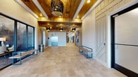 Located at 2480 Cherry Laurel Dr in Sanford, Florida, this single-story community clubhouse offers a thoughtfully designed gathering space with upscale finishes and a cohesive contemporary aesthetic. The grand entry lobby welcomes visitors with warm wood-paneled ceilings, a striking sculptural chandelier, an elevator, and large-format neutral tile flooring — setting an inviting tone from the moment of arrival. Seating benches line the corridor, guiding guests deeper into the facility. The central lounge area serves as the heart of the space, featuring a dramatic floor-to-ceiling white marble fireplace wall with a linear electric fireplace, a wall-mounted television, and built-in shelving with vibrant teal accents. Oversized windows and glass doors flood the room with natural light and provide views of the outdoor area. Adjacent to the lounge is a well-appointed café and bar station, complete with a long white quartz countertop, bar-height seating, dark textured tile backsplash, beverage equipment, and dual wall-mounted screens. An industrial-style overhead garage door opens the bar to the exterior. Additional flexible rooms with glass-paneled doors offer private or semi-private gathering options. Throughout the space, light tile flooring, recessed lighting, and carefully curated artwork contribute to a polished, resort-inspired ambiance.