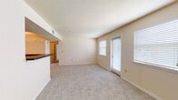 This single-floor residence offers a thoughtfully arranged layout that balances open communal spaces with private living areas. The living room serves as the centerpiece of the unit, featuring neutral-toned carpet, warm beige walls, and a textured ceiling. Multiple windows fitted with white horizontal blinds allow natural light to filter throughout the space, creating a bright and airy atmosphere. A door provides direct exterior access from the living area. A half-wall partition with a dark countertop separates the living room from the kitchen, creating a semi-open concept feel while maintaining a defined cooking zone.

The kitchen is equipped with rich medium-toned wood cabinetry, dark countertops, and black appliances, including an electric range and an over-the-range microwave. Light hardwood-style flooring grounds the kitchen, contrasting with the carpeted areas of the unit. Storage is well-represented through the generous upper and lower cabinet configuration.

The bedroom continues the neutral color palette with beige walls and plush carpet underfoot. A wide bifold closet provides ample storage, while a window brings in natural light. The overall design of the space is clean, functional, and move-in ready, offering a comfortable and practical living environment throughout.