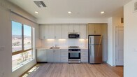 This contemporary single-floor unit is thoughtfully designed with a clean, modern aesthetic throughout. The kitchen serves as a standout feature, showcasing floor-to-ceiling cabinetry in a soft grey finish, a white subway tile backsplash, and a full suite of stainless steel appliances including a refrigerator, electric range, and over-the-range microwave. Light-toned countertops and an undermount sink complete the functional and stylish cooking space, which is bathed in natural light from a large adjacent window.

Light wood-tone flooring runs seamlessly throughout the unit, lending warmth and continuity to every room. The living area is generously sized, featuring large windows and a ceiling fan with a light fixture, creating a bright and airy atmosphere. Views of the surrounding community and distant mountain ridgelines are visible through the expansive glazing, adding a sense of openness to the space.

A sliding glass door provides direct access to an outdoor area, further extending the living space and enhancing natural ventilation. The bathroom is clean and well-appointed, featuring a warm-toned vanity with built-in drawers, a white countertop, a large mirror, and modern light bar fixtures. Recessed lighting throughout the unit adds a polished, contemporary finishing touch to this well-designed residence.