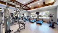 This single-level fitness center is thoughtfully designed to support a comprehensive workout experience. The space features a distinctive coffered ceiling with rich wood beam detailing and recessed lighting, lending the room a polished, upscale ambiance. Durable, light-toned concrete flooring spans the entire area, providing a practical and clean foundation throughout.

The cardio zone is equipped with multiple treadmills and an elliptical trainer, positioned along a windowed wall that offers natural light and views of an outdoor pool and surrounding landscape. A wall-mounted flat-screen television is positioned nearby for entertainment during workouts, and a standing floor fan provides additional ventilation.

The strength training area includes a multi-station cable machine, a seated weight machine, and an adjustable bench, complemented by a rack of graduated dumbbells. Large wall mirrors are strategically placed to maximize the sense of space and allow users to monitor their form. A rack of stability balls in multiple sizes and colors adds variety for functional fitness training.

Vibrant athletic wall art and subtle blue accent lighting contribute to an energizing atmosphere. The layout is open and well-organized, ensuring comfortable circulation between equipment zones throughout the facility.