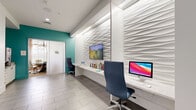 Located at 4460 Lower Park Rd in Orlando's Baldwin Park neighborhood, this single-floor commercial space combines contemporary design with a vibrant, welcoming atmosphere. The interior is defined by a crisp white palette accented by bold teal and orange color pops, creating a dynamic yet professional environment throughout.

The main lobby and common area features large-format gray tile flooring, recessed lighting, and a striking three-dimensional wave-textured accent wall that serves as a visual centerpiece. A built-in workstation along this wall is equipped with desktop computers and a wall-mounted display, providing a functional workspace within an elegant setting. Colorful curated artwork adds personality to the bright white walls.

A corridor connects the various areas of the space, distinguished by a vivid teal ceiling with pendant lighting that creates a memorable visual passage. Framed artwork lines the hallway walls, lending a gallery-like quality to the transition between spaces. Access-controlled doors lead to private offices or additional rooms along the corridor.

A dedicated conference room rounds out the space, featuring a round glass-top table surrounded by modern upholstered chairs, an artistic globe chandelier, a wall-mounted television, and sheer window treatments that allow abundant natural light to filter in. Together, these elements create a polished, design-forward commercial environment ideal for professional use.
