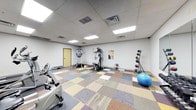 This single-floor fitness room offers a well-rounded workout environment designed to accommodate a variety of exercise routines. The space features a drop ceiling with recessed fluorescent panel lighting that ensures consistent, bright illumination throughout. The flooring consists of colorful, patchwork-style carpet tiles in muted earth tones, adding a casual and functional aesthetic underfoot.

The room is equipped with a multi-station cable weight machine positioned centrally, allowing for a range of strength training exercises. A dedicated medicine ball rack lines one wall, holding an assortment of weighted balls for functional training. A large stability ball rests nearby, suitable for core and balance work. On the opposite side of the room, a stationary recumbent bike provides cardiovascular training options. Free weights and additional small equipment are also visible, contributing to the room's versatility.

The neutral gray walls keep the space feeling open and clean, while a standard door provides access to the room. Ventilation is handled via ceiling-mounted air ducts, supporting a comfortable workout environment. Overall, the space is practical and efficiently organized for general fitness use.