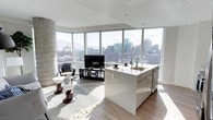 This 2-bedroom, 2-bathroom residence is thoughtfully designed with a modern, open-concept layout that maximizes both space and natural light. Expansive floor-to-ceiling windows wrap around the living and kitchen areas, flooding the interior with sunlight and offering commanding panoramic views of the city skyline—a standout feature that defines the ambiance of the entire unit.

The kitchen is sleek and contemporary, anchored by a large central island with a white high-gloss finish, an integrated sink, and stainless steel appliances, including a dishwasher. The clean lines and minimalist cabinetry create a polished aesthetic that flows seamlessly into the open living area.

The living space is warm and inviting, with light hardwood-style flooring, a distinctive curved concrete column accent, and generous room for comfortable seating and entertainment. The layout promotes a natural flow between the kitchen, dining, and living zones, making it ideal for both everyday living and hosting.

Throughout, the design palette is neutral and refined—white walls, light flooring, and modern finishes combine to create a bright, airy atmosphere. The sweeping city views visible from nearly every angle of the main living area serve as a constant, dramatic backdrop to this well-appointed urban residence.