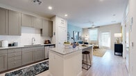 Located in Surprise, AZ, this well-appointed one-bedroom, one-bathroom residence offers a smart and efficient single-floor layout with modern finishes throughout. The open-concept kitchen and living area serves as the heart of the home, anchored by a granite-topped island with bar seating — ideal for casual dining or entertaining. The kitchen is outfitted with gray shaker-style cabinetry, white subway tile backsplash, stainless steel appliances including a refrigerator, range, microwave, and dishwasher, all set atop light wood-style flooring that extends throughout the main living areas. The living room features a soothing blue accent wall, ceiling fan, and large windows with drapes that invite abundant natural light. The bedroom continues the calming color palette with a teal accent wall, large window, and plush carpeting, offering a serene retreat. A dedicated walk-in closet with built-in shelving and hanging rods provides ample storage. The bathroom features a gray vanity with granite countertop, a large mirror, and light wood-style tile flooring. Recessed lighting and a clean, neutral color scheme tie the entire space together with a cohesive, contemporary aesthetic.