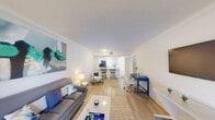 This single-floor residence offers a well-designed open-concept layout that seamlessly connects the main living areas. The kitchen is a functional centerpiece, featuring crisp white cabinetry, light quartz countertops, stainless steel appliances including a refrigerator, microwave, and dishwasher, and a peninsula breakfast bar with barstool seating. The kitchen opens directly into the living area, which is bright and inviting, benefiting from natural light that streams through large sliding glass doors leading to a private patio with lush outdoor views. Crown molding accents the living room walls, adding a refined architectural detail throughout. Light wood-look flooring runs through the main living spaces, contributing to the overall airy feel of the unit. The bedroom is a calm retreat, featuring light walls, wood-look flooring, and ample space for furnishings. The bathroom includes a white vanity with sink, a shower-tub combination, and tile flooring. An entry area with a closet and tile flooring connects the main spaces. Throughout the unit, a neutral white and light grey palette creates a clean, cohesive aesthetic, complemented by natural light from multiple windows.