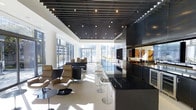 This sophisticated single-floor space showcases a striking contemporary design defined by a bold slatted ceiling in dark wood tones, punctuated by recessed lighting that creates a rhythmic visual pattern overhead. The open-plan layout seamlessly integrates multiple functional zones, beginning with a comfortable lounge area furnished with tan leather recliner chairs and low-profile dark seating arrangements. A long bar counter with sleek chrome stools runs centrally through the space, finished in polished black surfaces that reflect the abundant natural light. To one side, a well-appointed kitchen or service area features dark cabinetry, built-in appliances, and dark stone countertops, offering a refined and functional workspace. Floor-to-ceiling glazing wraps the perimeter, inviting panoramic outdoor views and bathing the interior in generous daylight. The light-toned tile flooring contrasts elegantly with the darker architectural elements throughout, enhancing the sense of spaciousness. The overall ambiance is one of modern luxury and clean sophistication, with a careful balance of dark and light tones that creates a polished, professional atmosphere throughout.