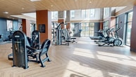 Beck Fitness is an impressive 3,100 square foot fitness facility designed to cater to a variety of exercise needs. The space is characterized by its soaring ceilings and expansive windows that flood the interior with natural light, creating an invigorating atmosphere for workouts. The flooring throughout is a stylish herringbone pattern in light wood tones, providing both aesthetic appeal and functionality.

The main area houses an extensive collection of cardio machines, including treadmills, ellipticals, and stationary bikes, strategically positioned to take advantage of the outdoor views. A dedicated strength training zone features a comprehensive array of weight machines and free weights. The layout is open and spacious, allowing for easy navigation between different workout stations.

Architectural elements such as exposed brick columns and a mix of warm and cool-toned walls add visual interest and define different functional areas. The facility also includes a separate studio space with mirrored walls, likely for group classes or personal training sessions. Modern amenities are evident throughout, including ceiling fans, recessed lighting, and what appears to be a refreshment or reception area with built-in cabinetry and tiled backsplash.

Overall, Beck Fitness presents a well-equipped, thoughtfully designed space that combines functionality with contemporary aesthetics to create an inviting environment for fitness enthusiasts of all levels.