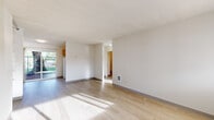 This single-floor unit is designed with a clean, functional layout that makes efficient use of its space. The main living area is finished with light-toned laminate wood flooring that runs throughout, complemented by neutral white walls and a textured ceiling. Natural light filters in through multiple windows, casting bright patterns across the floors and creating an open, airy atmosphere. A doorway connects the living area to additional rooms, maintaining a straightforward flow throughout the unit.

A dedicated entry area features a front door flanked by a window offering exterior views, alongside a full wall of built-in open shelving — a practical storage solution integrated directly into the architecture.

The kitchen is equipped with warm-toned wood cabinetry, white appliances including a refrigerator, electric range, and dishwasher, along with a ventilation hood. A sliding glass door in the kitchen provides direct access to an outdoor area and allows additional natural light to enter.

The bedrooms are carpeted in a neutral gray-beige tone and each include windows with exterior views and wall-mounted heating units. The bathroom features a combined tub and shower with a tiled surround, a grab bar, and light-toned vinyl flooring, providing a clean and practical space.
