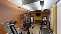 This fitness center offers a comprehensive workout environment designed to support a wide range of exercise routines. The space features warm terracotta-toned walls that lend an energizing ambiance, complemented by a light-colored ceiling with recessed panels and exposed industrial-style ductwork that adds a modern edge to the overall aesthetic.

The facility is outfitted with a variety of cardio equipment, including treadmills positioned along the perimeter, providing ample space for users to exercise comfortably. A multi-station cable and weight machine anchors the strength training area, offering versatility for full-body workouts. A dedicated free weights section lines one wall, featuring a tiered dumbbell rack stocked with a range of weights to accommodate all fitness levels. A large wall-mounted mirror adjacent to the free weight area enhances the sense of space and allows for proper form checks during exercise.

A flat-screen television is mounted on the wall, adding an entertainment element to keep users engaged during workouts. Overhead, linear pendant lighting provides bright, even illumination throughout the room. The flooring is a neutral, low-pile carpet suited for the equipment layout. Overall, the fitness center is thoughtfully arranged to maximize functionality while maintaining a welcoming and motivating atmosphere.