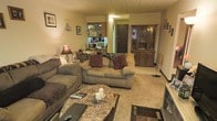 This single-floor space centers around a comfortable living area anchored by two tan upholstered sofas adorned with burgundy and brown accent pillows. A marble-topped coffee table serves as a focal point, complemented by a coordinating area rug that defines the seating zone. A classic wood china cabinet with glass-front upper doors provides elegant display and storage along one wall, while framed artwork and decorative wall sconces add character to the neutral-toned walls. A table lamp with a warm-toned shade contributes to the cozy ambiance throughout the space. An open passthrough or entryway connects the living area to what appears to be a kitchen beyond, where cabinetry and countertops are partially visible, creating a sense of continuity between spaces. The flooring transitions from carpeted living areas to what appears to be a hard surface near the entry. A solid front door provides the main point of entry. The overall atmosphere is warm and lived-in, with a traditional aesthetic reinforced by the wood furnishings, soft textiles, and layered decorative accessories throughout.