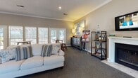 Located at 9989 Dorchester Rd in Summerville, South Carolina, this single-floor community clubhouse offers a thoughtfully designed gathering space that balances comfort and functionality. The open-concept layout is anchored by a spacious lounge area furnished with plush cream sofas, decorative throw pillows, and an abstract wall canvas, all set atop rich, textured carpet tiles. Crown molding runs throughout, lending a refined architectural detail to the warm, neutral-toned walls.

Natural light flows generously through large windows fitted with horizontal blinds, while a set of classic French doors opens directly to an outdoor pool and pergola area, seamlessly connecting interior and exterior living. A café-style high-top table with bar stools provides an additional seating nook, ideal for casual conversation or informal meetings.

Toward the rear of the space, a service counter with bar seating and granite-style surfaces serves as a welcoming reception or refreshment station, complete with a coffee machine and bar accessories. Adjacent to the counter, a dedicated workstation with a computer and printer offers a convenient business center for residents. Hardwood-style flooring in this zone contrasts nicely with the carpeted lounge, creating a natural visual separation of areas. Together, these features make the space a functional and inviting community hub.
