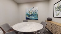 Located at 5550 Bingle Rd in Houston, TX, this conference room offers a refined and professional environment designed for productive gatherings. The centerpiece of the room is a striking round marble-top table, its white and grey veining adding a touch of elegance, surrounded by comfortable upholstered grey chairs. A bold, large-scale abstract painting in tones of teal, navy, and gold commands attention on the neutral-toned wall, infusing the space with a creative energy. The room is accented by a patterned carpet that complements the overall aesthetic. The conference room accommodates a maximum occupancy of nine and is accessed through a glass-paneled door that offers a view into the broader facility. The adjacent common area features light hardwood flooring, exposed beam ceilings, and eclectic décor including a live-edge wood piece and geometric tile accents, suggesting a stylish and well-appointed surrounding complex. Additional touches throughout the space include a framed mirror, a decorative wooden credenza with botanical artwork, and carefully chosen accessories, all contributing to a cohesive, modern-yet-warm atmosphere suited for professional use.