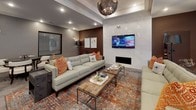 This well-appointed club room offers a thoughtfully designed, single-floor communal space divided into several distinct functional zones, each finished with modern sophistication. Large-format stone-look tile flooring flows throughout, anchoring the open layout and lending a cohesive, upscale feel.

The lounge area serves as the social centerpiece, furnished with tufted sofas accented by vibrant orange patterned pillows atop a richly colored area rug. A sleek linear fireplace set into a polished marble-style surround draws the eye, while a wall-mounted flat-screen TV above it adds entertainment value. A statement chandelier with an artisan metal frame hangs overhead within a tray ceiling detail, adding refined character to the space.

Adjacent to the lounge, a casual dining area features a live-edge wood high-top table with upholstered bar-height chairs positioned in front of dual wall-mounted screens—ideal for group viewing or social gatherings. Floor-to-ceiling glass entry doors flood this zone with natural light and provide views to the exterior.

A separate round table with elegant white chairs offers a more intimate dining or meeting setting, while recessed lighting throughout ensures the entire space is well-illuminated. Curated wall art and decorative floor lanterns add finishing touches that elevate the overall ambiance of this versatile, community-oriented space.