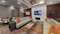 This well-appointed club room offers a thoughtfully designed, single-floor communal space divided into several distinct functional zones, each finished with modern sophistication. Large-format stone-look tile flooring flows throughout, anchoring the open layout and lending a cohesive, upscale feel.

The lounge area serves as the social centerpiece, furnished with tufted sofas accented by vibrant orange patterned pillows atop a richly colored area rug. A sleek linear fireplace set into a polished marble-style surround draws the eye, while a wall-mounted flat-screen TV above it adds entertainment value. A statement chandelier with an artisan metal frame hangs overhead within a tray ceiling detail, adding refined character to the space.

Adjacent to the lounge, a casual dining area features a live-edge wood high-top table with upholstered bar-height chairs positioned in front of dual wall-mounted screens—ideal for group viewing or social gatherings. Floor-to-ceiling glass entry doors flood this zone with natural light and provide views to the exterior.

A separate round table with elegant white chairs offers a more intimate dining or meeting setting, while recessed lighting throughout ensures the entire space is well-illuminated. Curated wall art and decorative floor lanterns add finishing touches that elevate the overall ambiance of this versatile, community-oriented space.