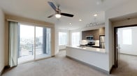 This single-floor space showcases a contemporary open-concept design that seamlessly connects the living area and kitchen. The living space is finished with plush neutral carpeting and features a ceiling fan with integrated lighting, complemented by recessed ceiling lights that create a bright, welcoming atmosphere. Large sliding glass doors lead to a private balcony, flooding the interior with generous natural light and offering views of the surrounding area.

The kitchen is a standout feature, boasting modern dark wood cabinetry with a warm grain finish, contrasted by a crisp white peninsula countertop that doubles as a breakfast bar. Stainless steel appliances, including a refrigerator, gas range, and microwave, are thoughtfully integrated into the design. Track lighting above the kitchen ensures excellent task illumination. The peninsula creates a natural boundary between the cooking and living spaces while maintaining the open, airy feel of the layout. Clean white walls and consistent modern finishes throughout contribute to a cohesive, contemporary aesthetic ideal for both everyday living and entertaining.