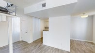 This two-floor space offers a bright and clean aesthetic defined by a neutral color palette and modern finishes. The main living area features wood-look plank flooring that adds warmth to the otherwise crisp, white interior. A structural half-wall elegantly separates the living area from the kitchen, creating a sense of division while maintaining an open, airy feel. The kitchen is equipped with white upper cabinetry and a dark contrasting countertop, providing a subtle visual contrast against the light surroundings. Vertical blind window treatments allow for flexible light control, while a central HVAC vent in the ceiling ensures year-round climate comfort. The textured ceiling adds a layer of dimension to the space. A solid entry door with a deadbolt provides a secure point of entry. The overall ambiance is minimalist and move-in ready, with a layout that lends itself well to functional everyday living. Spanning two floors, the space offers room for defined living and private areas, making it a practical and versatile environment suited to a variety of uses.