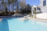 This inviting outdoor amenity space centers around a generously sized swimming pool with crystal-clear blue water, framed by a well-maintained concrete deck that provides ample room for lounging and socializing. The pool area is thoughtfully appointed with multiple seating arrangements, including cushioned chaise lounges, bar-height tables and chairs, and a shaded umbrella station, offering residents a variety of options for relaxation and outdoor entertaining.

A standout feature of the space is a pergola-covered lounge area outfitted with comfortable teal sectional seating accented by vibrant throw pillows, creating a cozy retreat with dappled natural light filtering through the overhead structure. This seating nook opens to sweeping views of the surrounding mature woodland, enhancing the sense of privacy and natural serenity.

The pool is enclosed by a sleek wrought-iron fence, ensuring security while maintaining open sightlines to the tree-lined landscape. A multi-story residential building with large windows and covered balconies serves as an attractive backdrop, adding architectural character to the setting. The combination of abundant natural light, lush greenery, and thoughtfully designed outdoor furnishings creates a resort-like atmosphere that is both functional and aesthetically appealing throughout the seasons.