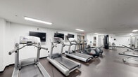 This fitness center offers a generously sized, single-level exercise room designed to support a comprehensive workout experience. The space is finished with durable, dark rubberized flooring throughout, providing a practical and resilient surface suited for a wide range of physical activities.

The cardio area is well-appointed with multiple treadmills and elliptical machines arranged in a row, each paired with a wall-mounted flat-screen television for entertainment during workouts. A stair climber and stationary bike round out the cardio offerings, ensuring a variety of aerobic options.

The strength training section features a selection of cable and weight machines, including a multi-functional cable tower and additional resistance equipment. A dedicated free weights area houses a full rack of dumbbells alongside a weight bench, supported by large wall mirrors that enhance the sense of space and allow users to monitor their form.

Exposed ventilation ductwork runs along the ceiling in the strength area, lending an industrial character to the otherwise clean, white-walled interior. Recessed and flush-mounted fluorescent lighting provides consistent, bright illumination throughout. A water fountain is conveniently accessible within the space. The overall layout is open and efficiently organized, accommodating both cardio and strength training needs within a single, functional facility.