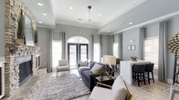 This well-appointed clubhouse offers a sophisticated and inviting environment designed for comfort and community. The main lounge area immediately draws attention with its elevated tray ceilings accented by crisp crown molding, recessed lighting, and a ceiling fan that adds both function and style. Soft gray walls create a calm, neutral backdrop that complements the modern yet comfortable furnishings throughout.

Natural light pours in through a series of large windows dressed with elegant floor-length drapes, as well as a striking arched transom window above double French doors that open to the exterior. This architectural detail serves as a focal point of the space, lending a classic charm to the otherwise contemporary interior. The French doors provide a seamless connection to the outdoors, enhancing the sense of openness.

The lounge is thoughtfully arranged with plush seating, including a deep charcoal sofa, accent chairs, and a layered area rug that anchors the space. A stone fireplace adds a warm, textural element to one side of the room. Adjacent to the lounge, a counter-height bar or dining area with seating offers a more casual gathering spot, ideal for socializing. The overall ambiance is polished, airy, and welcoming — a refined space suited for relaxation and community engagement.