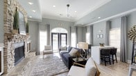 This well-appointed clubhouse offers a sophisticated and inviting environment designed for comfort and community. The main lounge area immediately draws attention with its elevated tray ceilings accented by crisp crown molding, recessed lighting, and a ceiling fan that adds both function and style. Soft gray walls create a calm, neutral backdrop that complements the modern yet comfortable furnishings throughout.

Natural light pours in through a series of large windows dressed with elegant floor-length drapes, as well as a striking arched transom window above double French doors that open to the exterior. This architectural detail serves as a focal point of the space, lending a classic charm to the otherwise contemporary interior. The French doors provide a seamless connection to the outdoors, enhancing the sense of openness.

The lounge is thoughtfully arranged with plush seating, including a deep charcoal sofa, accent chairs, and a layered area rug that anchors the space. A stone fireplace adds a warm, textural element to one side of the room. Adjacent to the lounge, a counter-height bar or dining area with seating offers a more casual gathering spot, ideal for socializing. The overall ambiance is polished, airy, and welcoming — a refined space suited for relaxation and community engagement.