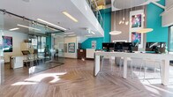 This contemporary single-floor commercial space is designed with a clean, open-concept layout that promotes both collaboration and functionality. The interior is immediately distinguished by striking teal accent walls that contrast sharply with crisp white surfaces, dark feature walls, and warm herringbone-patterned wood-look flooring that runs throughout the entire space.

The main area features a central workstation bar equipped with multiple computer monitors and modern bar-height seating, illuminated by a cluster of pendant lights and large disc-shaped hanging fixtures that descend from double-height ceilings. This setup provides a practical, tech-forward hub for client or staff interaction.

A glass-walled private office or meeting area sits to one side, enclosed by full-height frameless glass panels and sliding doors, offering visual transparency while maintaining acoustic separation. Inside, additional workstations and seating are arranged for consultations or focused work.

A comfortable lounge area features sleek, contemporary gray sofas arranged around polished round side tables, bathed in natural light streaming through expansive floor-to-ceiling windows that offer views of the surrounding urban streetscape. A large-scale colorful art piece adorns the teal wall nearby, adding a vibrant focal point.

Overall, the space conveys a professional yet welcoming ambiance, combining modern design elements, generous natural light, and thoughtfully curated furnishings.