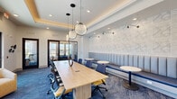 Located in Huntsville, Alabama, this Tech Hub is a thoughtfully designed coworking and collaboration space that balances professional functionality with contemporary style. The open floor plan centers around a large light wood conference table accommodating multiple occupants, surrounded by ergonomic blue and yellow task chairs. Above, a trio of striking spherical pendant lights hang from a coffered ceiling with warm cove lighting, lending the space a polished, modern ambiance. Along one wall, a continuous upholstered banquette in soft blue provides casual seating alongside round bistro tables, backed by a textured geometric accent wall fitted with swing-arm sconce lighting. A flat-screen display and credenza anchor the opposite end of the room for presentations. Glass-paned interior doors lead to private workrooms, offering quiet individual or small-group options. Large floor-to-ceiling windows and glass exterior doors flood the space with natural light while framing views of an outdoor courtyard and surrounding landscape. A welcoming lounge area near the entrance features plush gold accent chairs. Blue geometric-patterned carpet ties the design together throughout, creating a cohesive, vibrant workspace well-suited for meetings, focused work, and collaboration.