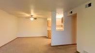 This single-floor space features an open-concept layout that connects the living area to the kitchen, creating a cohesive and functional flow throughout. The living area is finished with neutral wall-to-wall carpeting and crisp white walls, lending a clean and welcoming atmosphere. A ceiling fan with an integrated light fixture serves as a practical focal point overhead, providing both ambient lighting and air circulation.

A structural column partially separates the living space from the kitchen, adding architectural definition without fully closing off the two areas. The kitchen is equipped with light wood cabinetry that runs along the upper and lower walls, offering ample storage. Countertops feature a granite-style finish, adding a touch of visual interest. A pass-through window or opening in the dividing wall enhances the sense of openness and connectivity between the kitchen and living areas.

Tile flooring in the kitchen contrasts with the carpeting in the living area, clearly delineating each zone. HVAC vents are visible in the ceiling, and the overall neutral color palette throughout contributes to a bright, airy ambiance well-suited for a variety of furnishing styles.