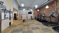 This fitness center combines industrial character with modern functionality, creating an inviting and well-appointed workout environment. The space is anchored by a dramatic exposed brick wall that adds texture and warmth, complemented by crisp white walls, exposed ductwork, and pendant-style industrial lighting that hangs from high ceilings throughout.

The cardio zone features a row of Landice treadmills, each equipped with individual monitors, positioned in front of a wall-mounted flat-screen television for entertainment during workouts. A built-in water station with a bottle-filling fountain provides convenient hydration access nearby.

The strength training area offers a barbell squat rack with an adjustable bench, a dual cable functional trainer, and a TRX suspension training system mounted to the wall alongside a detailed exercise reference chart. A rack of medicine balls rounds out the functional fitness options.

Patterned modular flooring in tones of gray and natural wood runs throughout, providing both durability and visual appeal. The entrance features warm wood-toned double doors, and a decorative vintage-style beer poster adds a playful, neighborhood nod to the overall aesthetic. The open single-floor layout ensures all equipment is easily accessible, making this fitness center a comprehensive space for a full range of training activities.