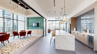 This elegantly designed amenity lounge occupies a generous single-floor footprint and is defined by its dramatic floor-to-ceiling curtain wall windows, which flood the interior with natural light and frame breathtaking panoramic views of the surrounding city skyline. Large-format slate-toned tile flooring runs throughout, providing a sleek, cohesive foundation for the space's varied functional zones.

The open-concept layout begins with a comfortable lounge area anchored by a tufted cognac leather sofa, complemented by contemporary upholstered chairs and a statement sputnik chandelier overhead. A wall-mounted flat-screen TV serves this seating zone, which opens directly to an outdoor terrace through expansive glass doors.

Adjacent to the lounge, a fully equipped kitchen features warm wood cabinetry, a striking iridescent blue zellige tile backsplash with brass fixtures, a stainless steel refrigerator, and an oversized marble-veined quartz island with bar seating beneath sculptural pendant lights.

A separate conference-style room, accessible via sliding glass doors, offers a distinctive design with bold teal zellige-tiled accent walls, a wall-mounted TV with open shelving, terrazzo-patterned carpeting, and generous window views. Additional casual dining tables with seating are positioned throughout to accommodate a variety of gathering needs. The overall ambiance is modern, refined, and welcoming.