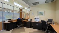 This single-floor office space is designed to support a functional and professional working environment. The workspace is anchored by a row of built-in dark-finished workstations equipped with desktop computers, printers, and office supplies, providing an organized setup for multiple users. Ample under-counter cabinetry with clean, simple hardware offers generous storage, keeping the work surfaces tidy and efficient.

A defining architectural feature is the floor-to-ceiling glazed partition wall with warm wood-toned framing, which separates the main workspace from an adjacent area while maintaining visual openness. Clerestory windows positioned above the partition allow natural light to filter in from above, complementing the recessed ceiling lighting and track lighting system overhead. The drop ceiling with integrated HVAC venting contributes to a polished, professional finish.

The neutral wall tones are accented by a framed landscape artwork, adding a touch of warmth to the otherwise utilitarian setting. Light-colored carpet flooring grounds the space and contributes to a quiet, focused atmosphere. Overall, the office balances practicality with a welcoming, well-lit ambiance suited to daily professional use.